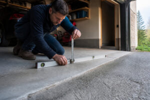Un homme accroupi dans son garage utilise un niveau à bulle et une règle pour mesurer la pente du sol.