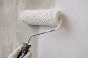 Une main gantée utilisant un rouleau à peinture pour appliquer une couche de peinture blanche sur un mur en béton gris.